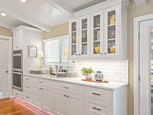 Kitchen featuring double oven, beamed ceiling, backsplash, white cabinetry, and glass insert cabinets