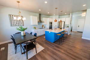 Kitchen with wall chimney range hood, appliances with stainless steel finishes, a chandelier, white cabinets, and an island with sink