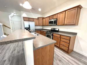 Kitchen with stainless steel appliances, brown cabinetry, a kitchen island with sink, light wood-style flooring, and dark countertops