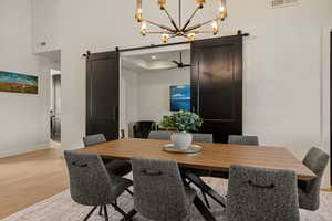 Dining area with a barn door, light wood-type flooring, a chandelier, ceiling fan, and a towering ceiling