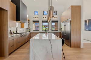 Kitchen with light stone countertops, light wood-style flooring, appliances with stainless steel finishes, a kitchen island with sink, and hanging light fixtures