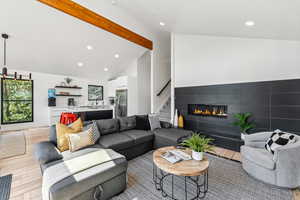 Living room with light wood finished floors, a tiled fireplace, beam ceiling, recessed lighting, and stairs