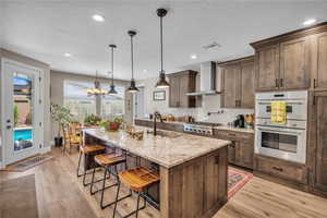 Kitchen featuring wall chimney exhaust hood, stainless steel appliances, light wood-style flooring, a kitchen bar, and light stone counters