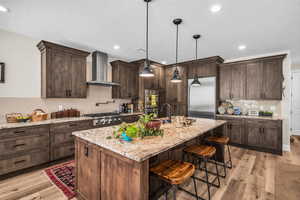 Kitchen with wall chimney range hood, appliances with stainless steel finishes, tasteful backsplash, dark brown cabinets, and a breakfast bar