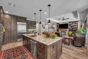 Kitchen with stainless steel appliances, ceiling fan, a stone fireplace, light wood-style floors, and light stone counters