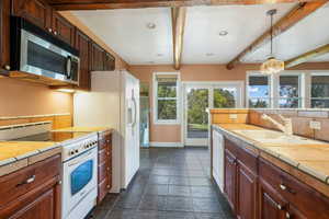 Kitchen featuring white appliances, tile countertops, beamed ceiling, decorative light fixtures, and dark tile patterned floors