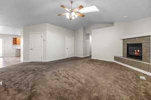 Unfurnished living room with lofted ceiling, dark carpet, a ceiling fan, a tiled fireplace, and a skylight