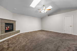 Unfurnished living room featuring vaulted ceiling, dark colored carpet, a tile fireplace, a skylight, and ceiling fan