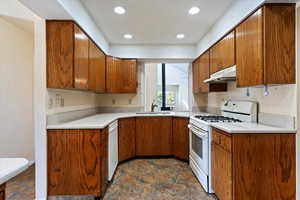 Kitchen with white appliances, brown cabinets, under cabinet range hood, light countertops, and recessed lighting