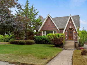 Tudor-style house with brick siding, a front lawn, an outdoor structure, and roof with shingles