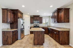 Kitchen with appliances with stainless steel finishes, a center island, dark brown cabinets, recessed lighting, and light stone counters