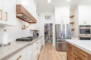 Kitchen with appliances with stainless steel finishes, open shelves, custom range hood, light wood finished floors, and white cabinetry
