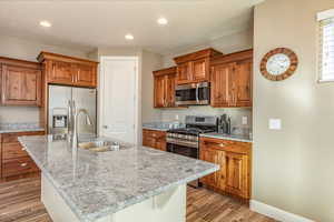 Kitchen with appliances with stainless steel finishes, brown cabinetry, light wood-type flooring, light stone counters, and recessed lighting