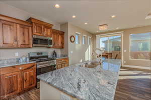 Kitchen with appliances with stainless steel finishes, brown cabinets, recessed lighting, dark wood-type flooring, and light stone countertops