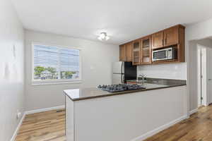 Kitchen featuring stainless steel microwave, black gas stovetop, brown cabinets, freestanding refrigerator, and light wood-style flooring