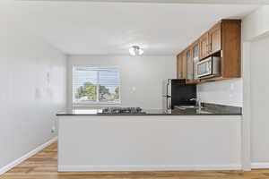 Kitchen with stainless steel microwave, gas stovetop, brown cabinetry, freestanding refrigerator, and dark stone counters