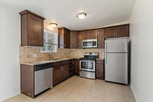 Kitchen with appliances with stainless steel finishes, backsplash, dark brown cabinetry, light stone counters, and light tile patterned floors