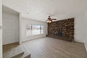 Unfurnished living room with ceiling fan, wood finished floors, a stone fireplace, and a textured ceiling