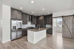Kitchen with appliances with stainless steel finishes, dark stone counters, tasteful backsplash, wall chimney exhaust hood, and light wood-style flooring