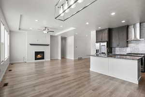 Kitchen with wall chimney exhaust hood, stainless steel appliances, tasteful backsplash, a tray ceiling, and a kitchen island with sink