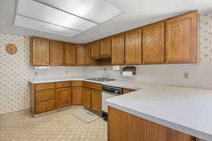 Kitchen with wallpapered walls, brown cabinetry, light countertops, dishwasher, and light flooring