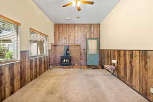 Living room featuring wood walls, a wood stove, wainscoting, carpet flooring, and ceiling fan