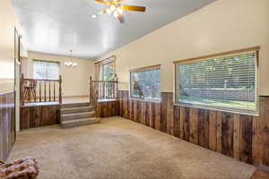 Carpeted empty room featuring wood walls, wainscoting, a chandelier, ceiling fan, and stairs