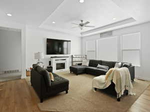 Living area with a raised ceiling, a ceiling fan, recessed lighting, wood finished floors, and a fireplace