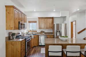 Kitchen featuring stainless steel appliances, dark wood-style floors, light stone counters, a breakfast bar, and brown cabinetry