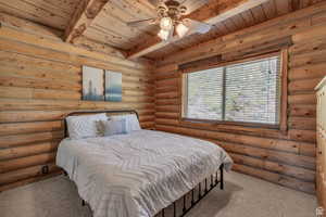 Carpeted bedroom featuring a wood ceiling with exposed beams, rustic walls, and ceiling fan