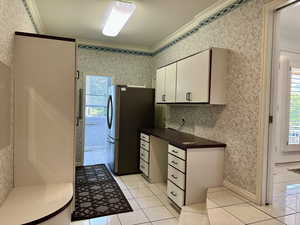 Pantry/storage area featuring crown molding, white cabinets, freestanding refrigerator, light tile patterned flooring, and wallpapered walls