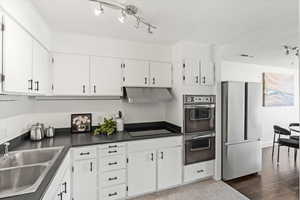 Kitchen with stainless steel fridge, track lighting, multiple ovens, dark countertops, and white cabinetry