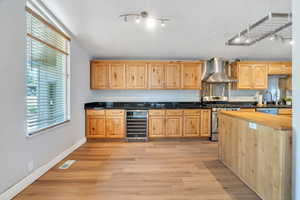 Kitchen with track lighting, beverage cooler, appliances with stainless steel finishes, wall chimney range hood, and light wood-style flooring
