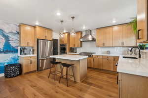 Kitchen featuring appliances with stainless steel finishes, wall chimney exhaust hood, light wood-type flooring, a kitchen bar, and a kitchen island