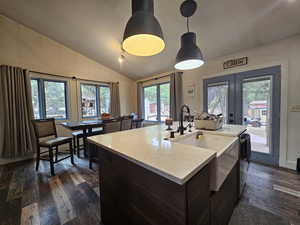 Kitchen featuring french doors, vaulted ceiling, dark wood-style floors, a center island with sink, and decorative light fixtures