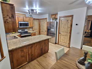Kitchen with stainless steel appliances, a peninsula, light wood-type flooring, light stone counters, and brown cabinetry