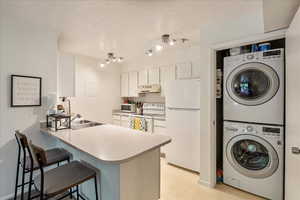 Kitchen with stacked washing machine and dryer, white appliances, a peninsula, white cabinets, and a kitchen bar