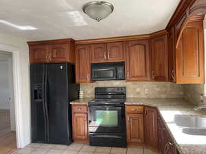 Kitchen with black appliances, decorative backsplash, light stone counters, light tile patterned floors, and brown cabinetry