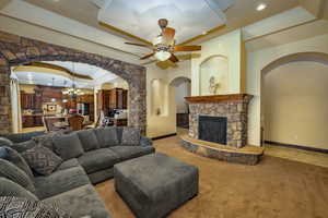 Living room featuring a tray ceiling, arched walkways, a chandelier, light colored carpet, and a stone fireplace