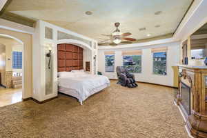 Carpeted bedroom with arched walkways, a raised ceiling, a glass covered fireplace, and ceiling fan