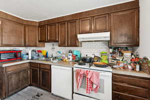 Kitchen with white appliances, under cabinet range hood, backsplash, light countertops, and a textured ceiling