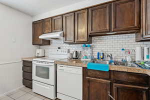 Kitchen featuring white appliances, dark brown cabinets, under cabinet range hood, backsplash, and a textured ceiling