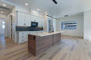 Kitchen with light wood-style floors, light countertops, an island with sink, backsplash, and white cabinetry