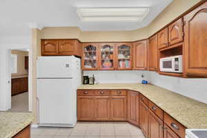 Kitchen with white appliances, light tile patterned flooring, brown cabinets, glass insert cabinets, and light stone countertops