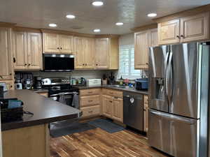 Kitchen featuring stainless steel appliances, dark countertops, light brown cabinets, dark wood-style flooring, and recessed lighting