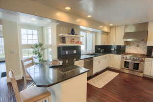 Kitchen featuring appliances with stainless steel finishes, a peninsula, wall chimney exhaust hood, open shelves, and decorative backsplash