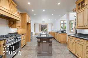 Kitchen featuring recessed lighting, stainless steel gas range, custom exhaust hood, dark stone counters, and a center island with sink