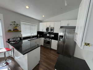 Kitchen featuring a peninsula, dark countertops, stainless steel appliances, vaulted ceiling, and white cabinetry