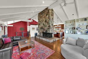 Living room featuring beamed ceiling, wood finished floors, high vaulted ceiling, a stone fireplace, and ceiling fan