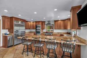 Kitchen with a peninsula, built in appliances, light stone countertops, wall chimney exhaust hood, and recessed lighting